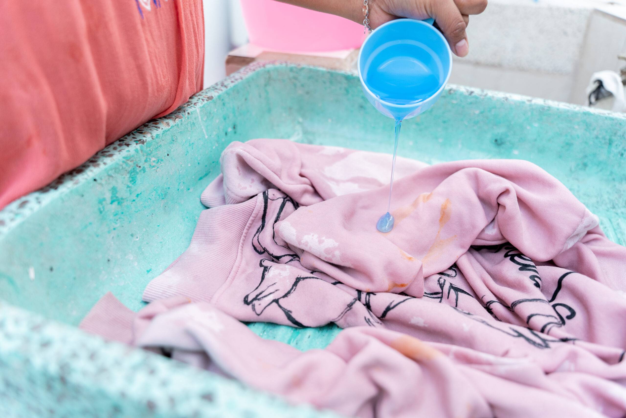 A young Hispanic woman is pouring liquid detergent on a pink sweatshirt to handwash on a traditional sink