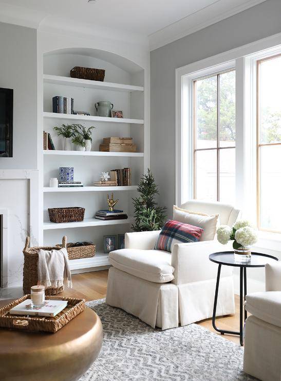 Arch built-in bookcase styles and displays decorative pieces, books and plants in a living room furnished with white skirted accent chairs and a round gold coffee table atop a gray wool rug.