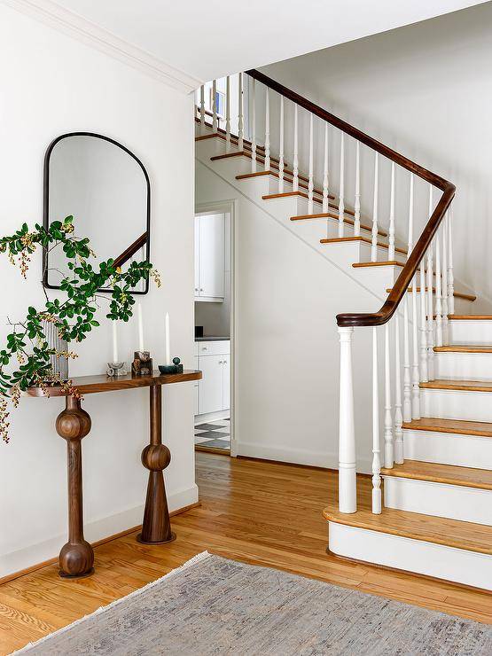 A black arch mirror hangs in a foyer over a brown oval console table.