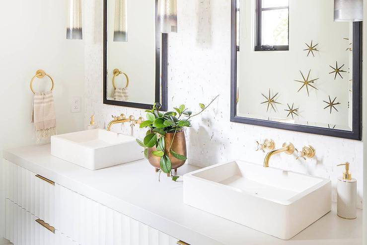 His and hers vessel sinks on a white rippled floating washstand with wall mount antique brass faucets and black beveled mirrors. Brass sunburst wall decor adds the perfect styled appeal for this transitional bathroom.