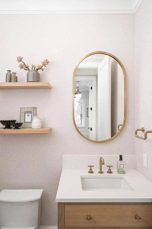 Powder room features a brass oblong mirror mounted on pink wallpaper over a brown wooden washstand with brass pulls and an aged brass gooseneck faucet and stacked shelves over the toilet.