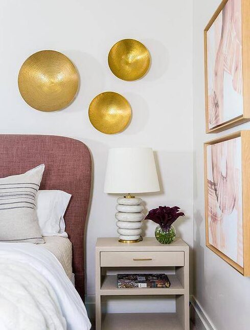 Chic bedroom updated with a mauve headboard and a light taupe nightstand displaying marble rings lamp under hammered brass wall decor. White and pink abstract wall art faces the bed with an artistic and flare complementing the rose tones in the mauve headboard.