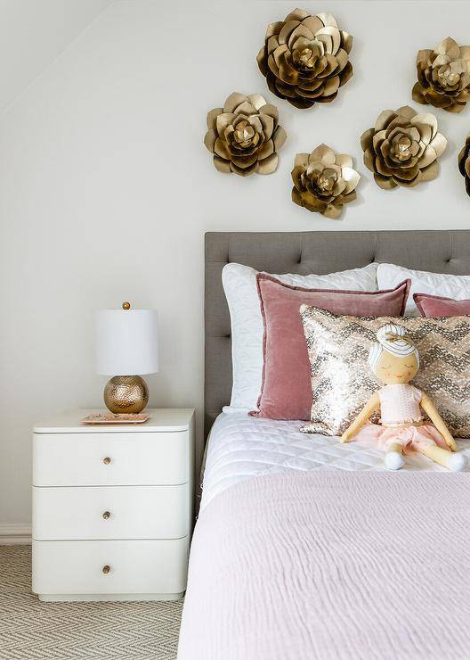 Pink and gray girl's room features a curved white nightstand accented with brass knobs and a hammered gold lamp and positioned beside a charcoal gray upholstered headboard. The headboard, located beneath gold lotus wall decor, supports a bed styled with pink velvet pillows layered behind a pink and gray chevron metallic pillow.