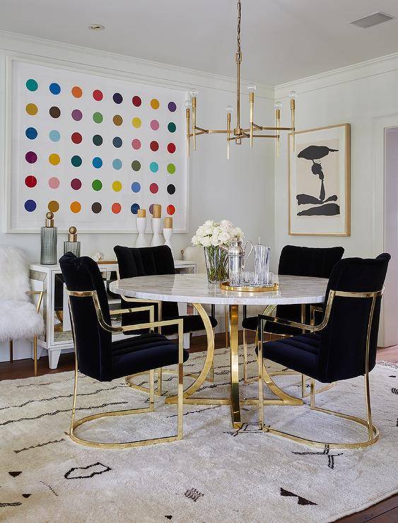 Black velvet and brass dining chairs join a round Capiz dining table illuminated by a brass 8 light chandelier in a contemporary dining room boasting various wall art and styled decor.