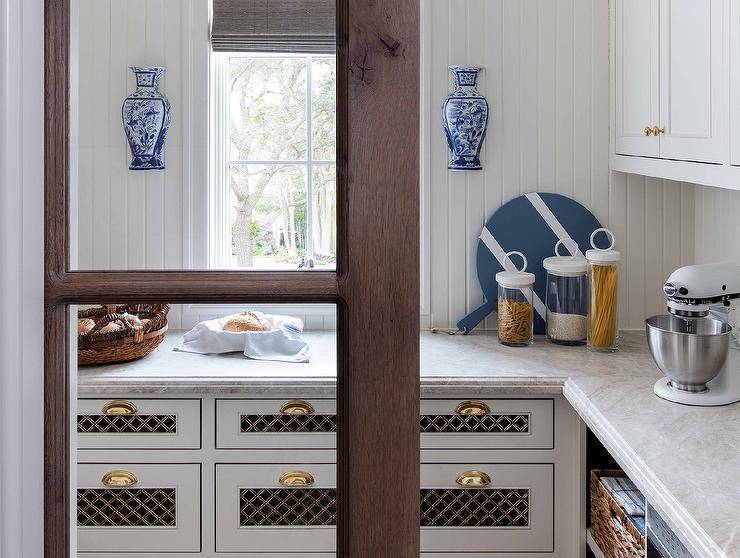 White and gold lattice pantry doors are complemented with brass cup pulls and a beveled marble countertop fixed against beadboard trim beneath a window located between chinoiserie wall decor.