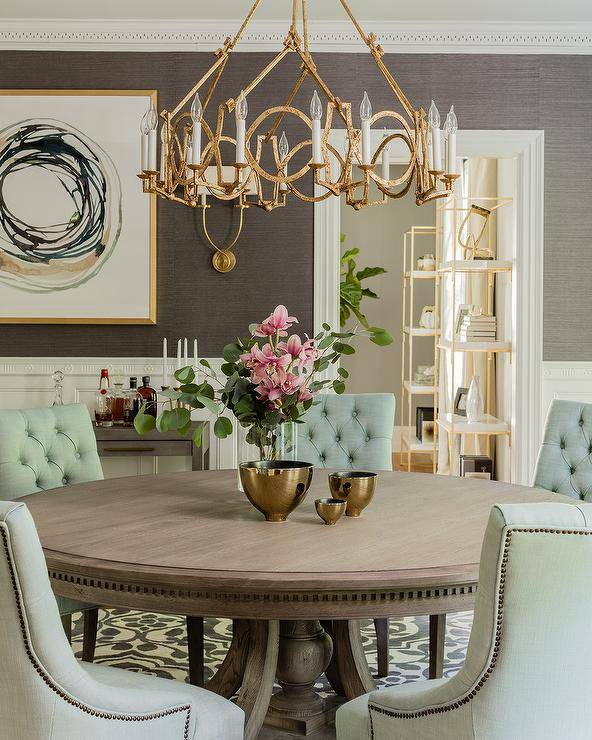 A gold leaf ring chandelier takes center stage over a round wood dining table surrounding green tufted linen chairs. Brass tableware, lovely floral centerpiece, and elegant background decor bring a transitional dining room design to life. Symmetric Twist Sconces next to a gold and black abstract wall art increases the room's ambience and attraction.