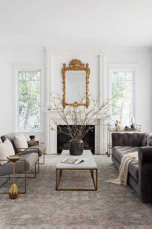 Elegant living room features a marble and brass coffee table placed on a gold and gray rug between side-by-side gray leather tufted accent chairs and a gray velvet chesterfield couch. An eye-catching gold baroque mirror hangs over a white fireplace mantel. A brass bar cart sits beside the fireplace.