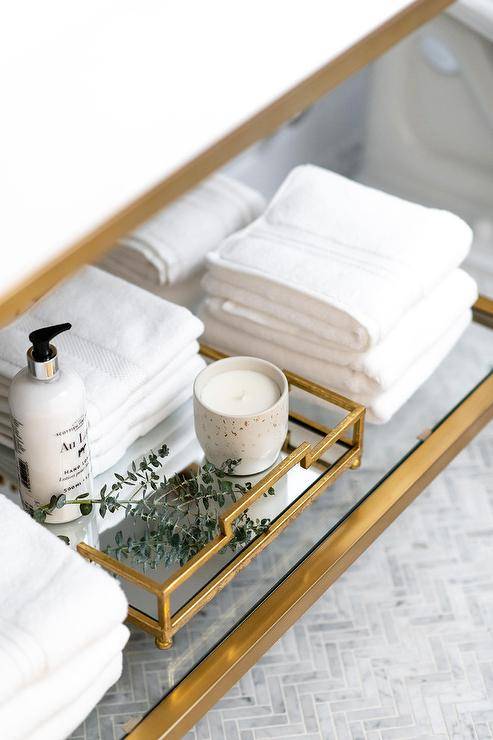 Brass washstand features a glass shelf styled with decor and stacked towels. White and gray marble herringbone floor tiles complete the bathroom with a classic material and contemporary style.