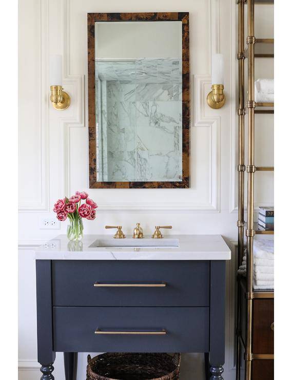 Transitional bathroom with a burl wood mirror flanked by brass sconces mounted on millwork walls above a black washstand topped with marble countertop and a brass vintage faucet. A brass etagere brings a French appeal to the bathroom displaying decor and bathroom essentials.