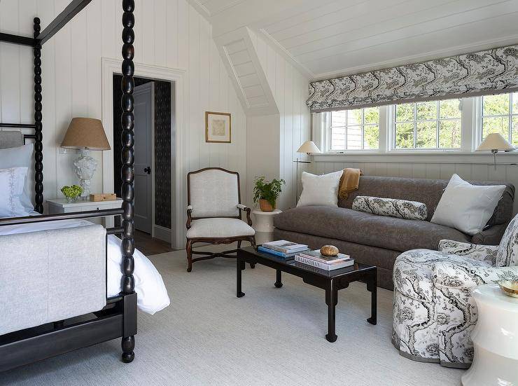 Cottagecore bedroom sitting area features a black chinoiserie coffee table placed on a gray rug in front of a gray damask couch topped with white pillows flanking a white and black pillows bolster pillow. The bolster pillow is made in fabric matching an upholstered accent chair and a white and black roman shade covering a row of windows flanked by polished nickel swing arm floor lamps. Walls and a vaulted ceiling are clad in shiplap trim.