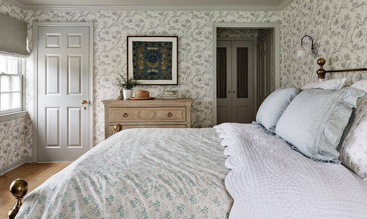 Charming cottage bedroom is clad in gray floral wallpaper lined with gray crown molding and baseboards. Art hangs beside a light gray door and over French beige dresser adorned with brass hardware.