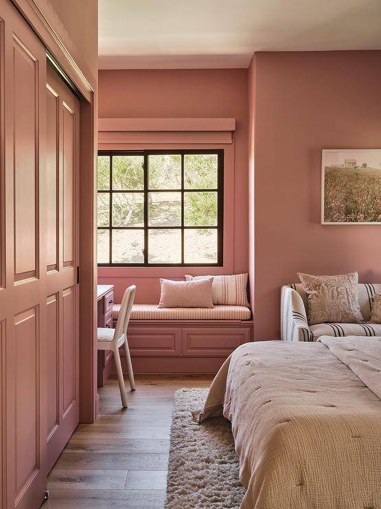 Mauve pink bedroom walls complement mauve pink sliding closet doors fixed beside a mauve pink desk positioned adjacent to a mauve pink built-in window seat. The window seat is mounted beneath a window covered in a mauve pink roman shade.