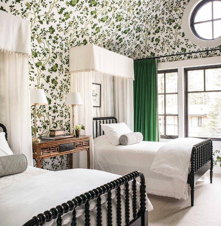Green botanical wallpaper covers the walls over a gorgeous bedroom featuring black spindle beds contrasted with white valances and white curtains. The beds are topped with black stripe bolster pillows and flank a brown bamboo nightstand accented with tall brass lamps. Windows are covered in green curtains.
