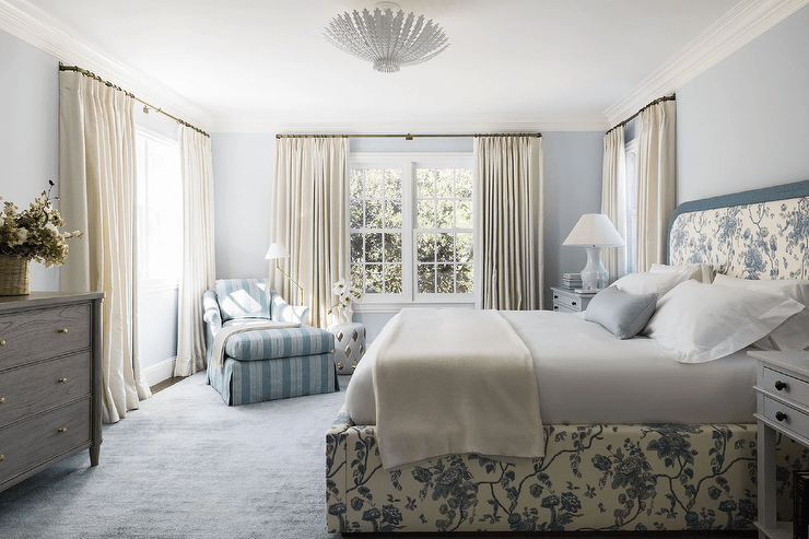 White and blue cottagecore bedroom boasts a brass floor lamp and a white lattice stool placed beside a blue striped chaise lounge positioned on a blue rug in a corner between windows dressed in ivory curtains. Pale blue walls are complemented with white crown moldings.