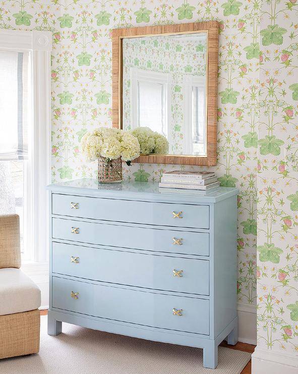 A raffia mirror hangs from a wall covered in Strawberry Trellis Wallpaper over a sky blue dresser in a cottagecore bedroom.