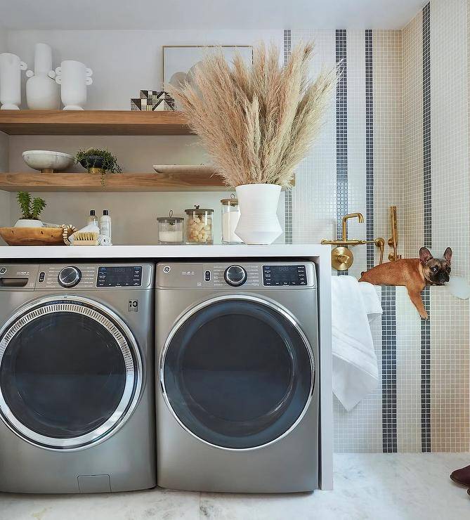 Wood floating shelves are stacked over an enclosed silver front loading washer and dryer located beside a white and gray striped grid tiled dog shower with an antiqute brass shower kit.