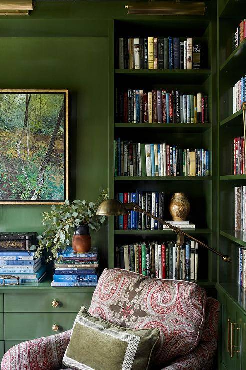 Welcoming home office features glossy green lacquered bookcases lit by brass picture lights and fitted with a brass swing arm sconce fixed above a pink and gray locked chair topped with a green velvet pillow. Art hangs from a green all over books stacked atop green drawers with brass pulls.