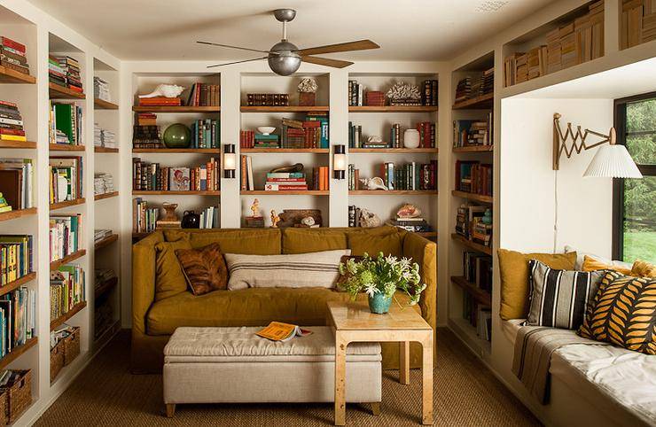 Cottage style home library boasts white and wooden built-in floor-to-ceiling bookshelves, a brown slipcovered sofa, and a built-in window seat lit by a brass accordion arm wall lamp.