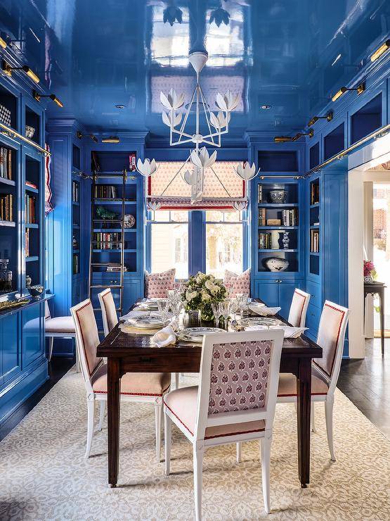 This elegant blue home library and dining room features a cream rug placed beneath a wooden dining table surrounded by block print French dining chairs lit by a white tulip chandelier hung from a blue lacquered ceiling. At the end of the table, a window dressed in a red grosgrain roman shade flanked by glossy blue built-in bookshelves lit by brass picture lights.