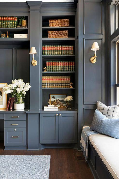 A dark gray built-in window seat is topped with a white cushion and fixed adjacent to a dark gray built-in bookcase flanked by brass sconces mounted to millwork accented gray walls. The shelves are located over gray shaker cabinets finished with brass knobs and located beside a built-in desk mounted beneath overhead bookshelves.
