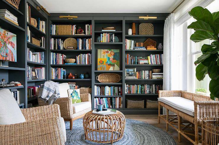Home library features black built in bookcase lit by brass picture lights, wicker accent chairs with a rattan coffee table atop a round blue jute rug and a wicker bench under a window with a tall potted plant.