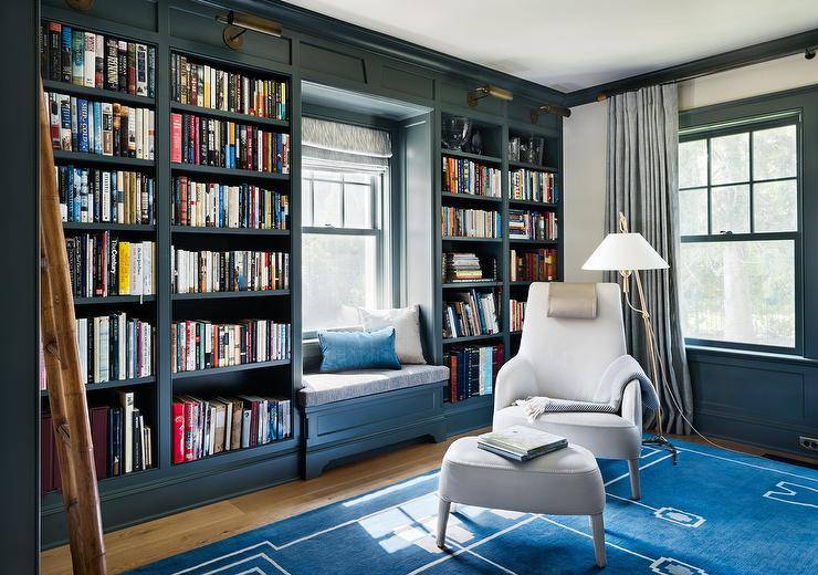 Dark blue paneled den boasts a white chair with matching ottoman placed atop a blue rug illuminated by a brass floor lamp. Home library features a wall of dark blue built-in bookshelves illuminated by antique brass picture lights flanking a dark blue built-in window seat bench.