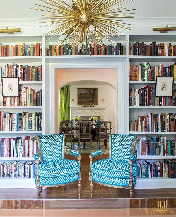 An eye-catching brass sputnik pendant hangs above two turquoise blue upholstered French chairs placed in front of a doorway framed by built in white bookshelves lit by the brass picture lights mounted above them.