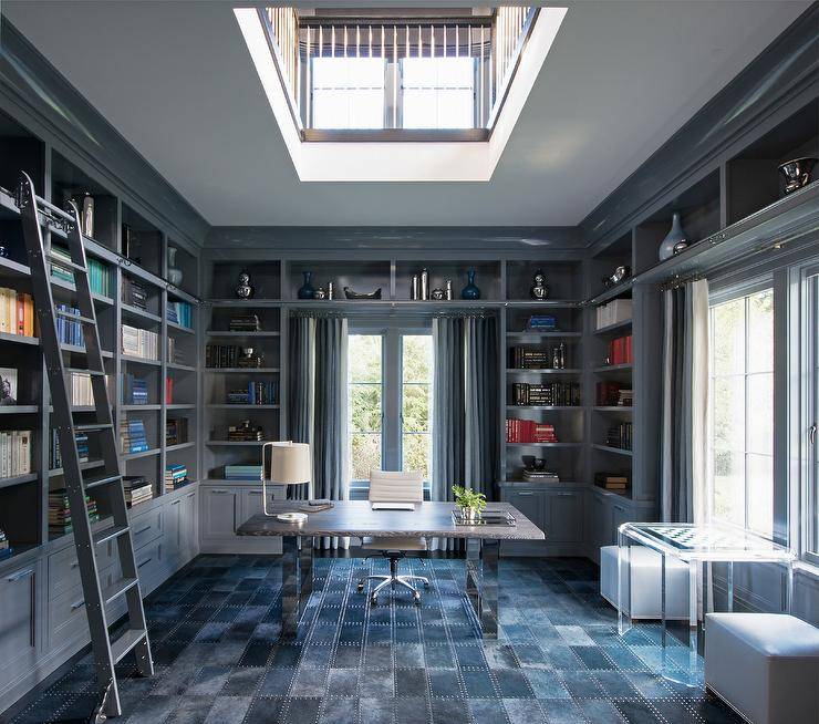 Stunning stately home features a two story library featuring walls fitted with gray built-in bookshelves fixed over gray cabinets and matched with a gray ladder. A window dressed in blue grommet curtains is positioned behind a stained gray oak and nickel desk seating a white leather task chair on a blue suede grid rug. Thick gray crown moldings line the walls, as an acrylic chess table is positioned beneath a window and between blue leather cube poufs.