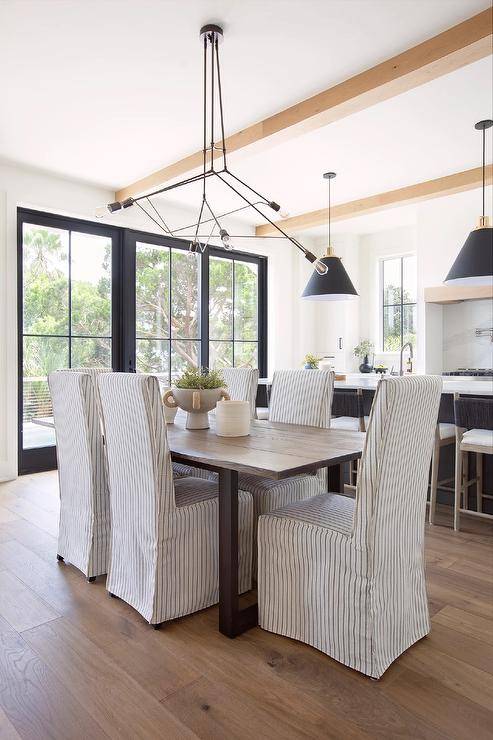 An oil rubbed bronze atom chandelier illuminates gray pinstripe slipcovered dining chairs placed around a wood and metal industrial dining table.