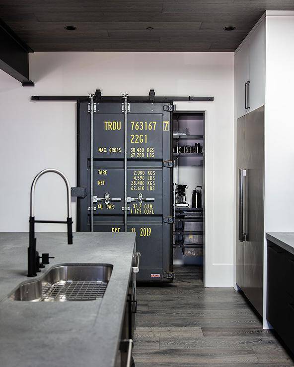 Under a brown plank ceiling, an industrial door on rails opens to a kitchen pantry.