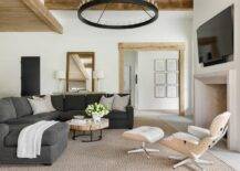 A black wagon wheel chandelier illuminates a wonderfully styled living boasting a round industrial coffee table placed on a bound sisal rug in front of a charcoal gray sectional complemented with light gray pillows.