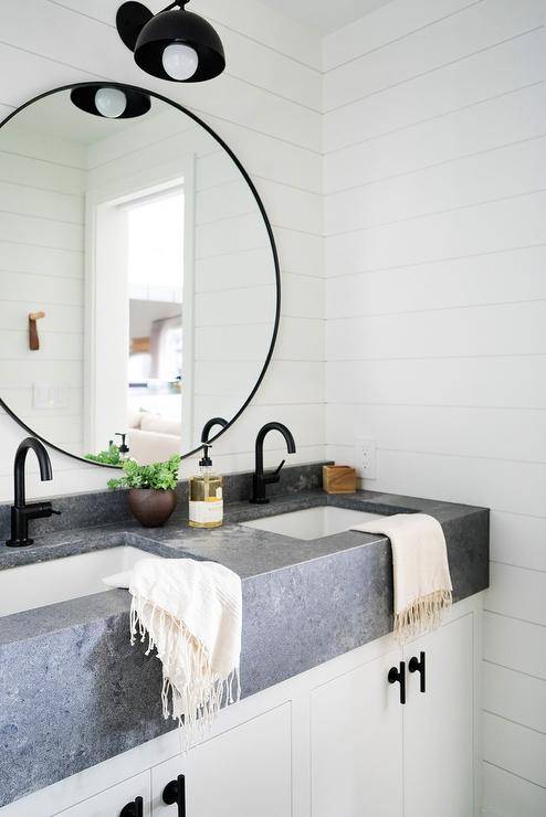Cottage bathroom with an industrial touch features a white washstand with a concrete countertop and oil rubbed bronze faucets. A shiplap wall sets a farm style surface for round black mirrors and a black farm style sconce.
