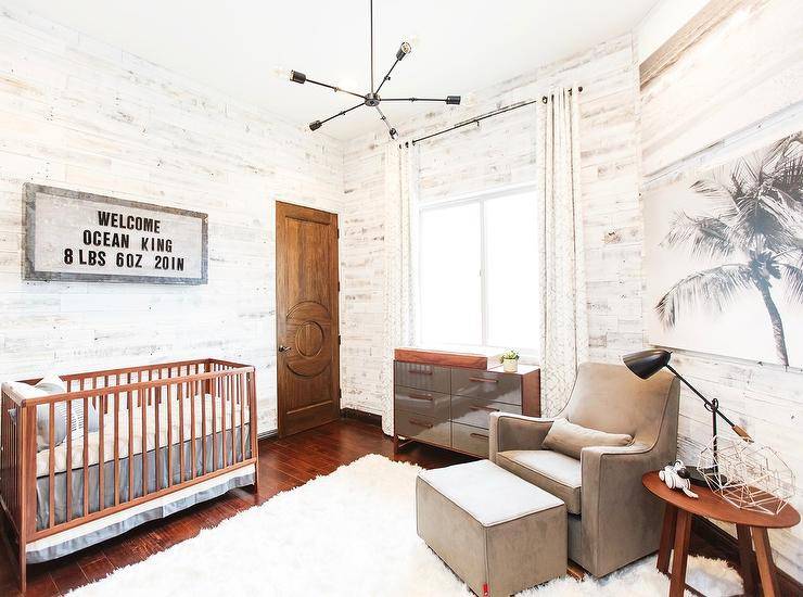 A wood birth announcement art over the brown crib makes the nursery a unique and modern space designed with a white wool rug, brown mirrored dresser and a gray nursery glider and ottoman. An Ellis adjustable arm chandelier further delivers an industrial touch lighting up the whitewashed plank walled room with a trendy fixture.