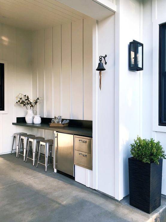 Covered outdoor kitchen features a black countertop bar with tolix stools and a stainless steel mini fridge. Concrete floors bring an industrial finish to the look balanced with white plank walls.