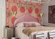 Luxurious eclectic bedroom features a pink velvet Moroccan style headboard placed against a Suzani blanket hung from a brass rod and lit by gold tiered pendants mounted over gray French nightstands. The bed is covered in beige bedding topped with a pink velvet lumbar pillow. Gray striped French chairs sit at the foot of the bed on a red and black rug.