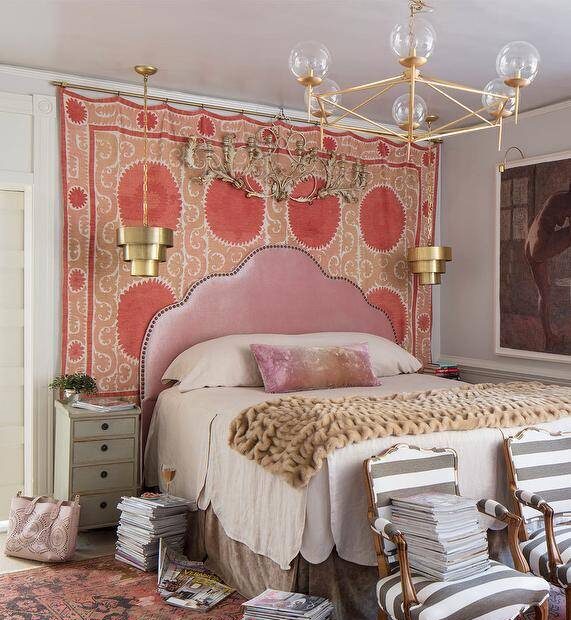 Luxurious eclectic bedroom features a pink velvet Moroccan style headboard placed against a Suzani blanket hung from a brass rod and lit by gold tiered pendants mounted over gray French nightstands. The bed is covered in beige bedding topped with a pink velvet lumbar pillow. Gray striped French chairs sit at the foot of the bed on a red and black rug.