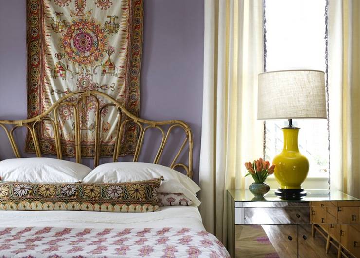 Gorgeous bedroom with lilac purple walls paint color, tapestry wall art, wicker headboard, mirrored nightstand and glossy yellow lamp.