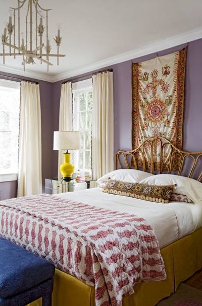 Chic, eclectic bedroom with lilac walls paint color, tapestry wall art, wicker headboard, yellow bed skirt, blue velvet bench, mirrored nightstand, glossy yellow lamps and white faux bamboo chandelier.