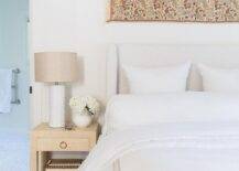This gorgeous bedroom features a restful ambiance starting with a white wingback headboard dressed in white and tan linen bedding. A gold grasscloth wrapped nightstand displays a white greek key lamp with surrounding accents from a long tapestry over headboard and white faux fur rug.