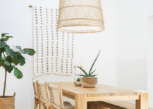 A large basket chandelier lights a chunky oak dining table paired with white leather and wooden dining chairs and positioned in front of a white and gray geometric tapestry hung from a white wall.