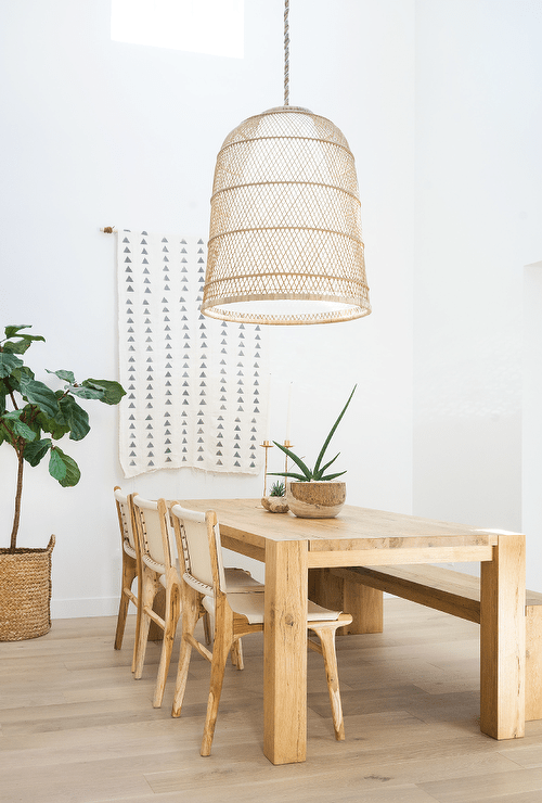 A large basket chandelier lights a c،ky oak dining table paired with white leather and wooden dining chairs and positioned in front of a white and gray geometric tapestry ،g from a white wall.