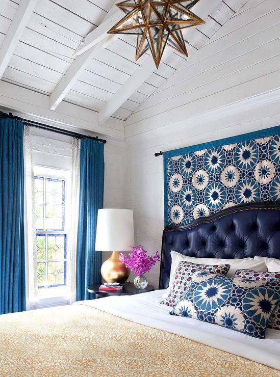 White plank vaulted ceiling holds a glass and brass Moravian star pendant over a bed dressed in a yellow medallion blanket accented with white sheets topped with a blue medallion pillow layered in front of blue and red print pillows and matching a tapestry hung behind the navy blue leather tufted headboard. Beside the bed, a round black beside table lit by a gold leaf table lamp positioned beside a window dressed in ocean blue linen curtains.