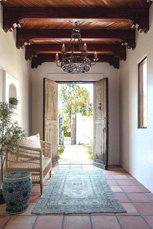 Mediterranean style foyer features a turquoise blue overdyed rug on a terracotta grid floor with a rattan bench and a reclaimed wood double front doors illuminated by a wrought iron chandelier hung from a ceiling with ornate ceiling beams.