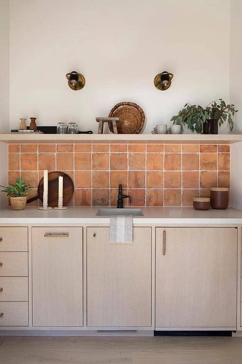 A gray wash oak floating shelf is mounted over terracotta grid backsplash tiles, while an oil rubbed bronze faucet is fixed over gray wash oak cabinets adorned with brass hardware.