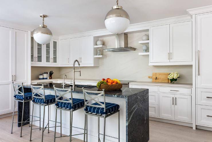 Black marble waterfall countertop on a white shaker kitchen island boasting iron counter stools with blue stripe cushions illuminated by nickel globe lights.