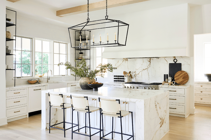 Darlana Linear Chandelier lights a white shaker island topped with a marble waterfall edge countertop seating ivory leather stools.