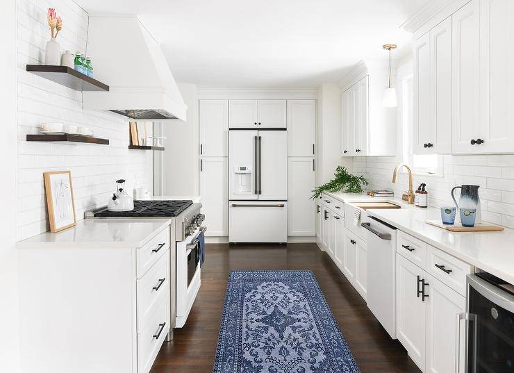 Classic white kitchen features white cabinets with black pulls surrounding a modern white refrigerator and a blue runner.