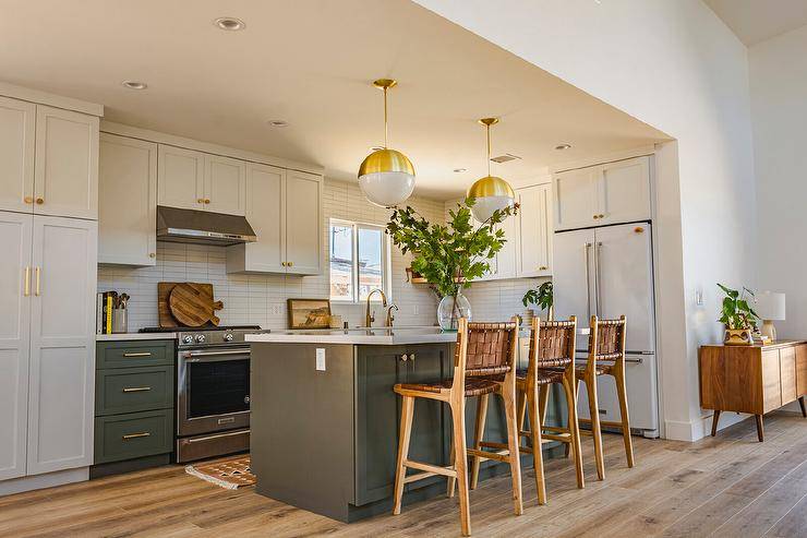 White and gray kitchen features white and gold globe pendants that illuminate a dark gray center island with brown woven leather stools, white cabinets with brass pulls and a white refrigerator.