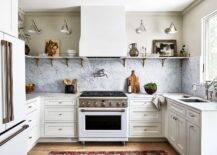 White and gray kitchen features long marble and brass shelves lit by a Boston functional library lights, a white hood over a nickel swing arm pot filler and white stove and marble backsplash, a white French door refrigerator and white cabinets.