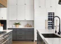 Well-appointed white and gray kitchen features dark gray lower cabinets accented with oil rubbed bronze pulls and a marble-like countertop fixed against a white exposed brick backsplash and beneath stacked white shaker upper cabinets finished with orb knobs. A hidden white shiplap fridge with a glass front door is inset beneath white shaker cabinets.